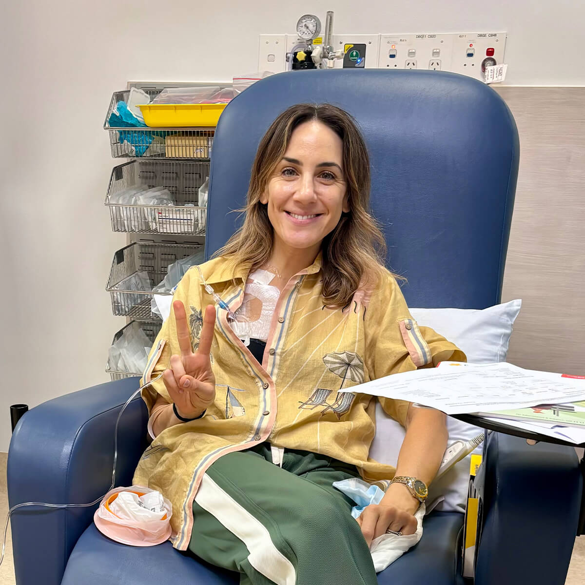 A woman in a hospital chair smiles and gives a peace sign, wearing a patterned shirt and green pants, with medical equipment nearby.