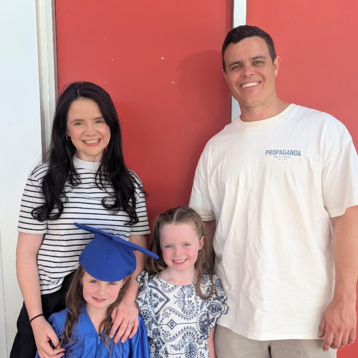 A family of four poses together; two girls in graduation attire, with parents smiling behind them against a red wall.