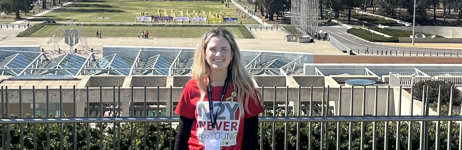 A smiling woman in a red shirt stands in front of a park with a green lawn and people in yellow shirts in the background.