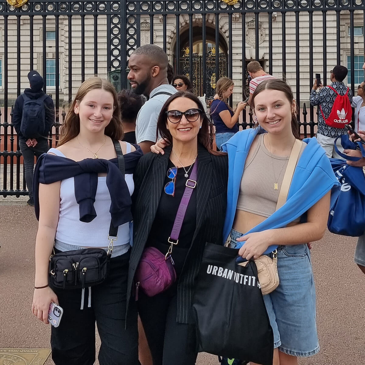 Three women pose in front of a gate, smiling. A crowd and buildings are visible in the background.