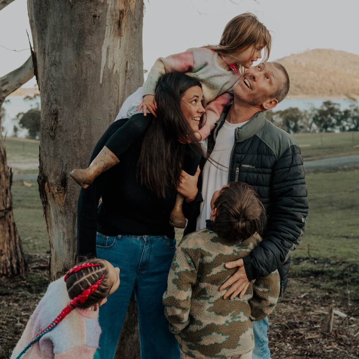 A family of four playfully interacts outdoors near a tree, with two children on their parents' shoulders.