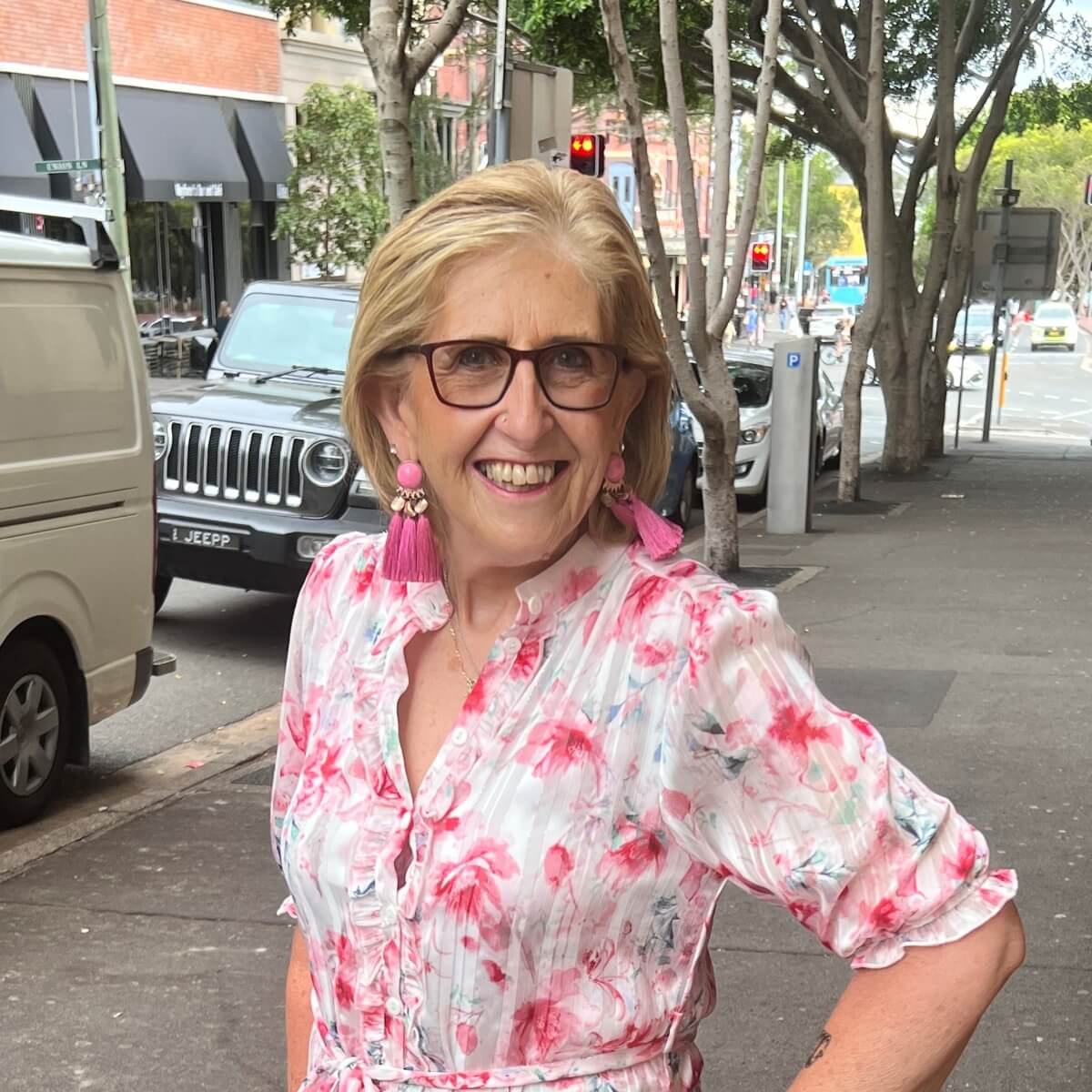 A smiling woman with glasses and pink tassel earrings stands on a city street, wearing a floral blouse.