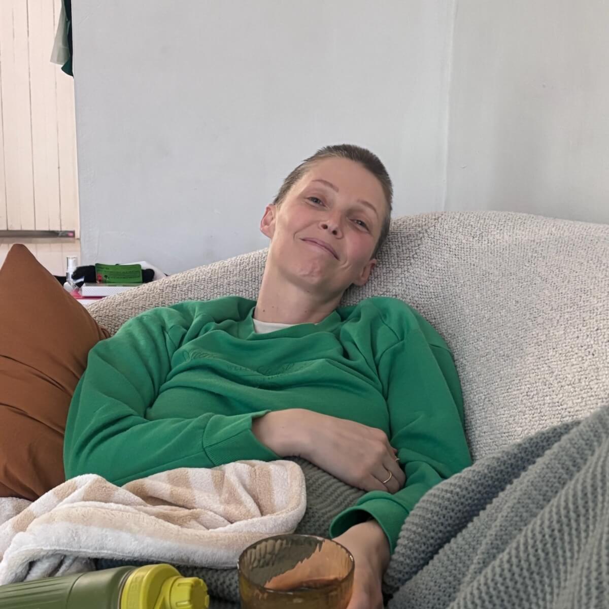 A person in a green sweatshirt sits on a couch, smiling, with a drink in hand and blankets around them.