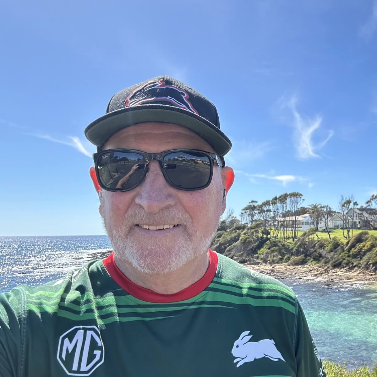 A man with glasses and a cap smiles for a selfie by the ocean, with trees and a house in the background.