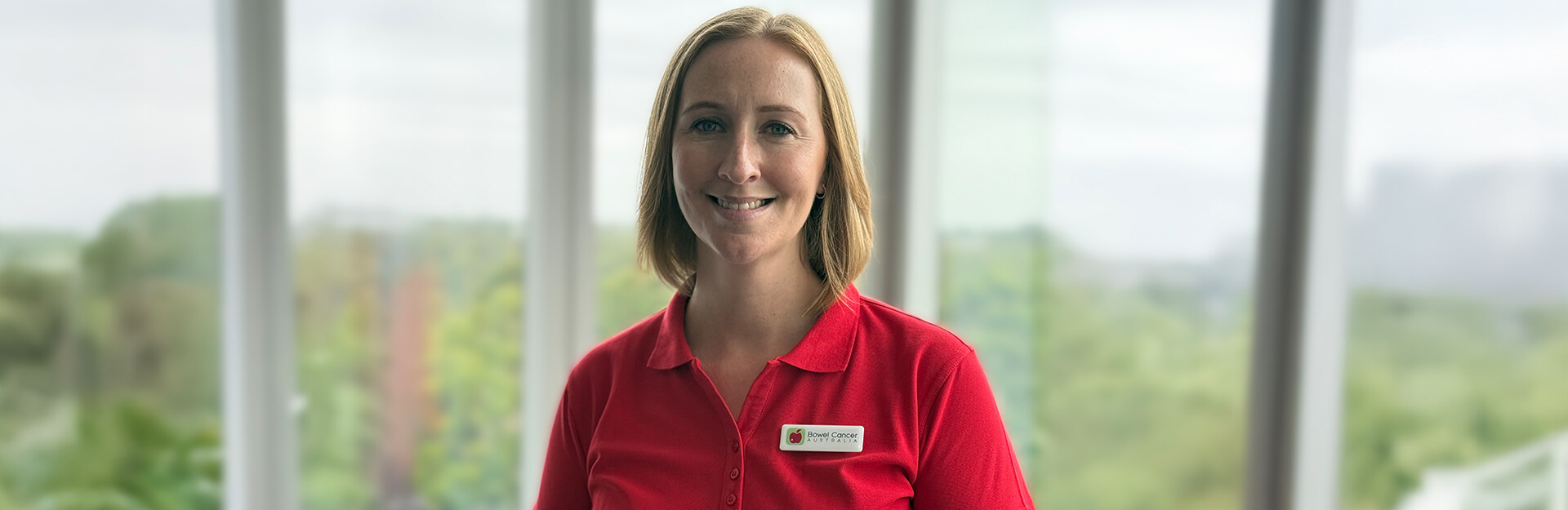 A smiling woman in a red shirt stands in front of large windows with a green landscape visible outside.
