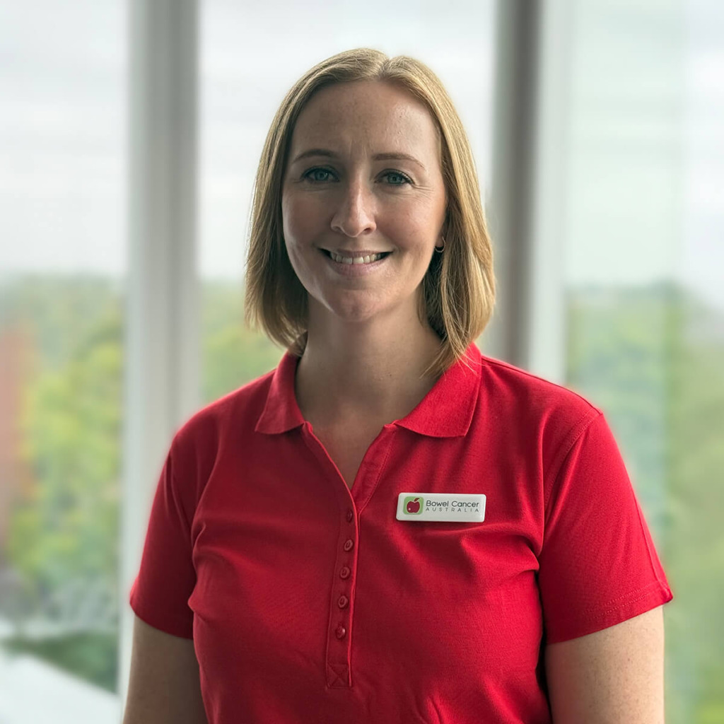 A woman with shoulder-length hair smiles, wearing a red polo shirt with a badge that says "Bowel Cancer Australia."