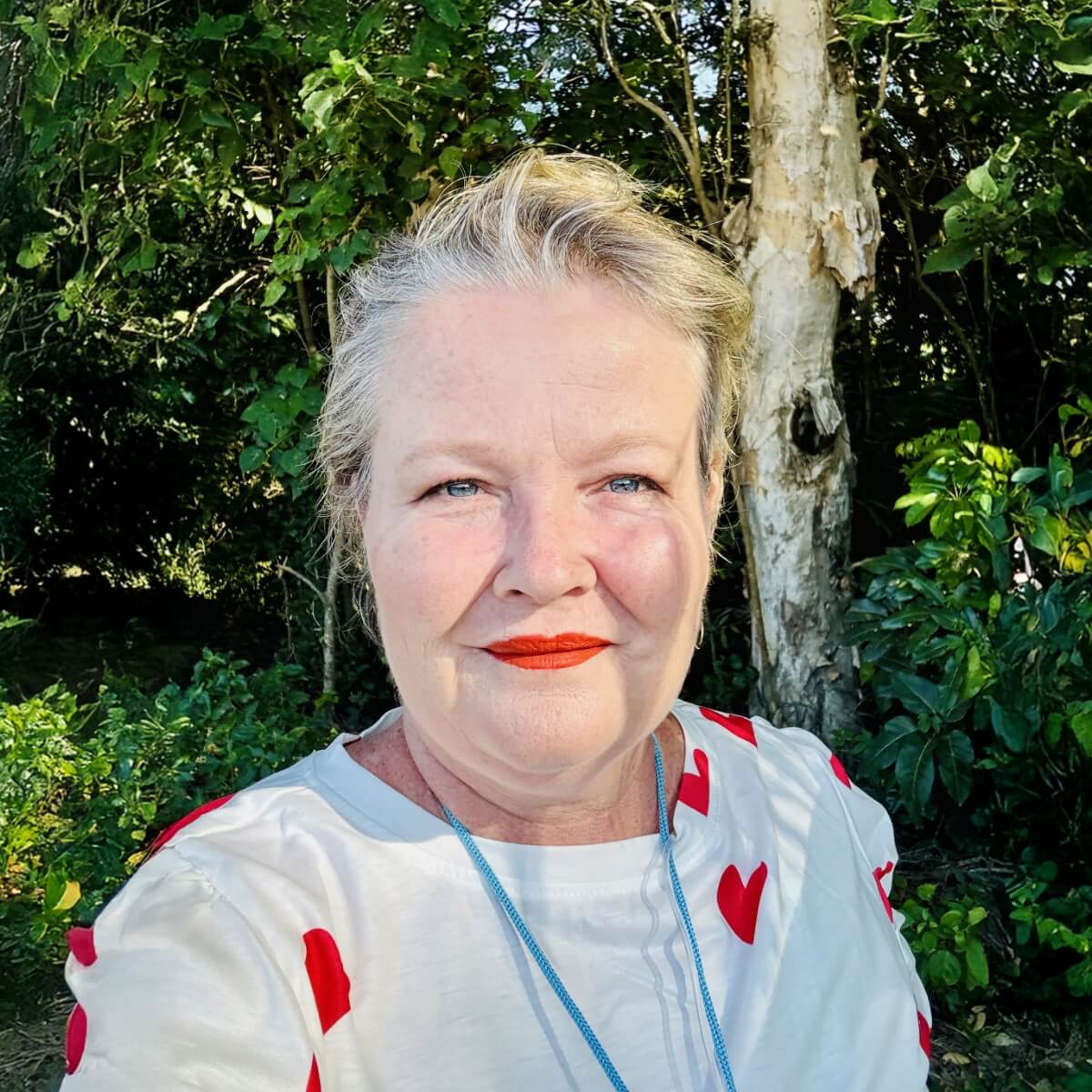 A woman with short hair, wearing a white shirt with red hearts, poses outdoors with greenery and a tree in the background.