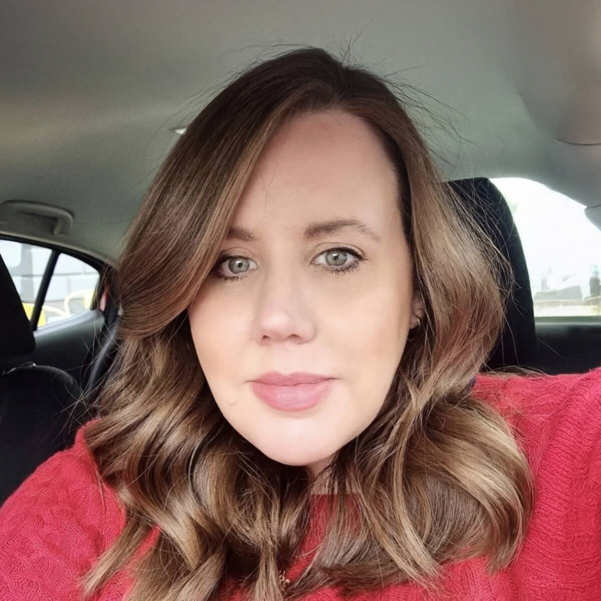 A woman with long, wavy brown hair wearing a red sweater, seated in a car, looking at the camera.