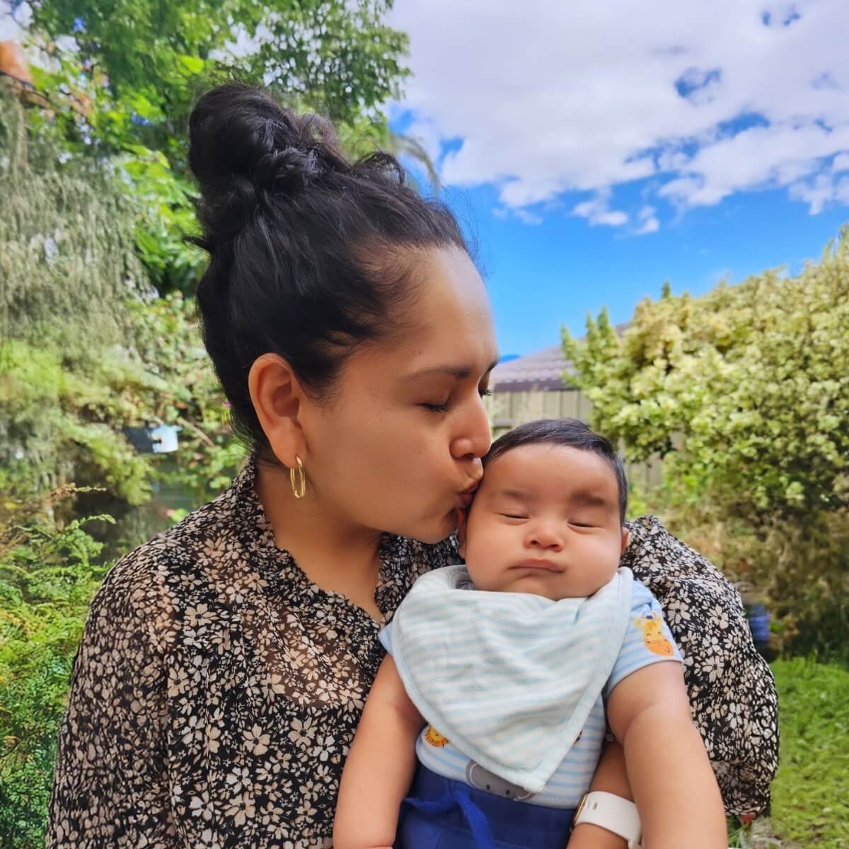 A woman kisses a baby in a garden, surrounded by greenery and a blue sky. The baby looks content.