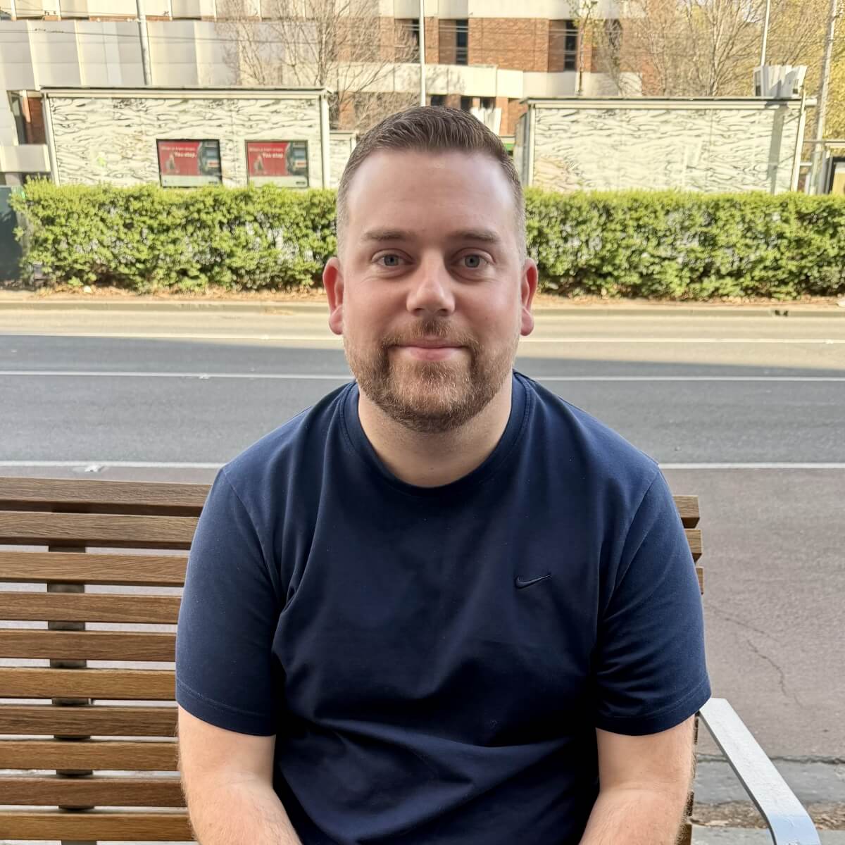 A man with a beard sits on a bench outdoors, wearing a navy blue shirt, with greenery and buildings in the background.