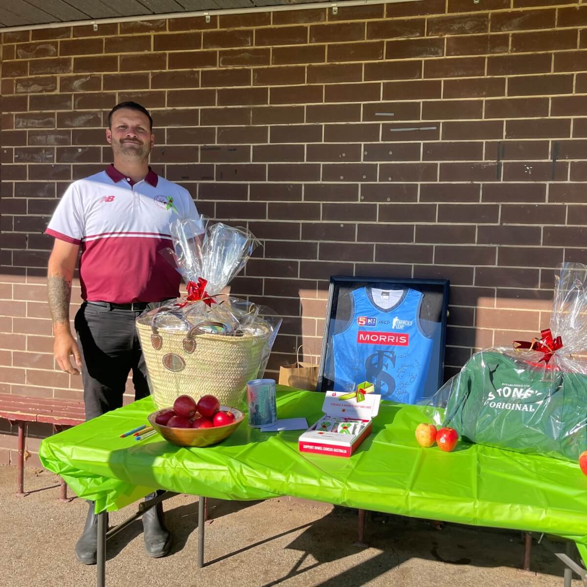 A man stands beside a table with a green tablecloth, featuring a gift basket, apples, a framed jersey, and a green shirt.