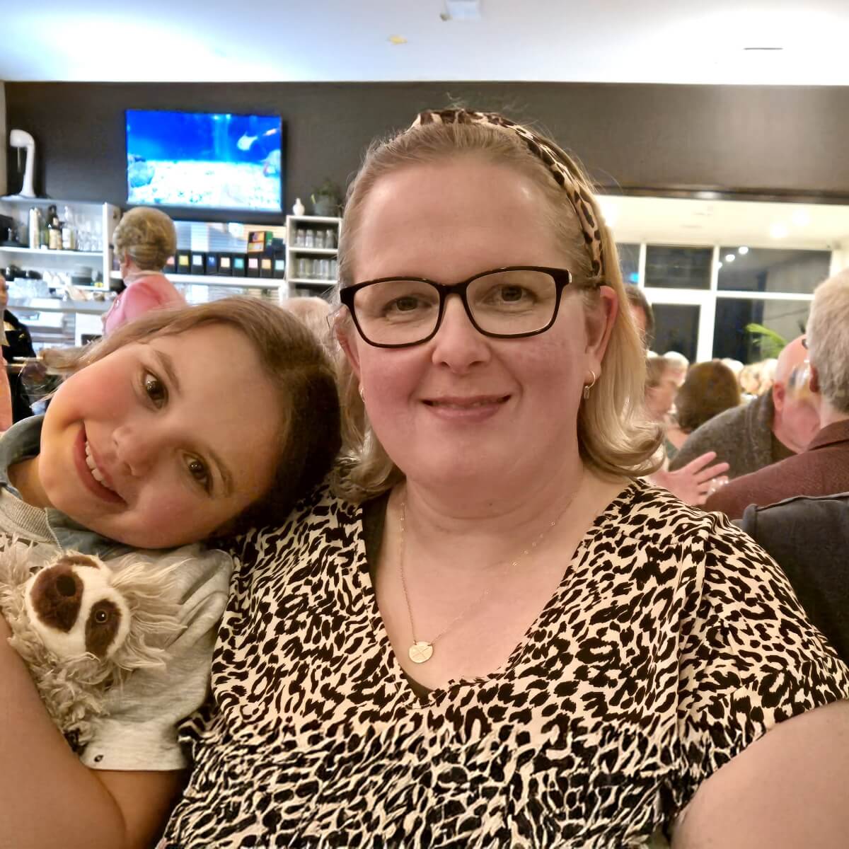 A woman and a girl smile together, with the girl holding a stuffed sloth. A lively restaurant scene is in the background.