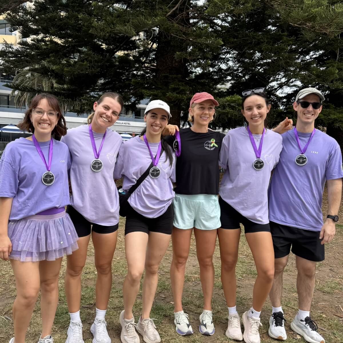 A group of six people wearing purple shirts and medals pose together outdoors, smiling in front of a tree.