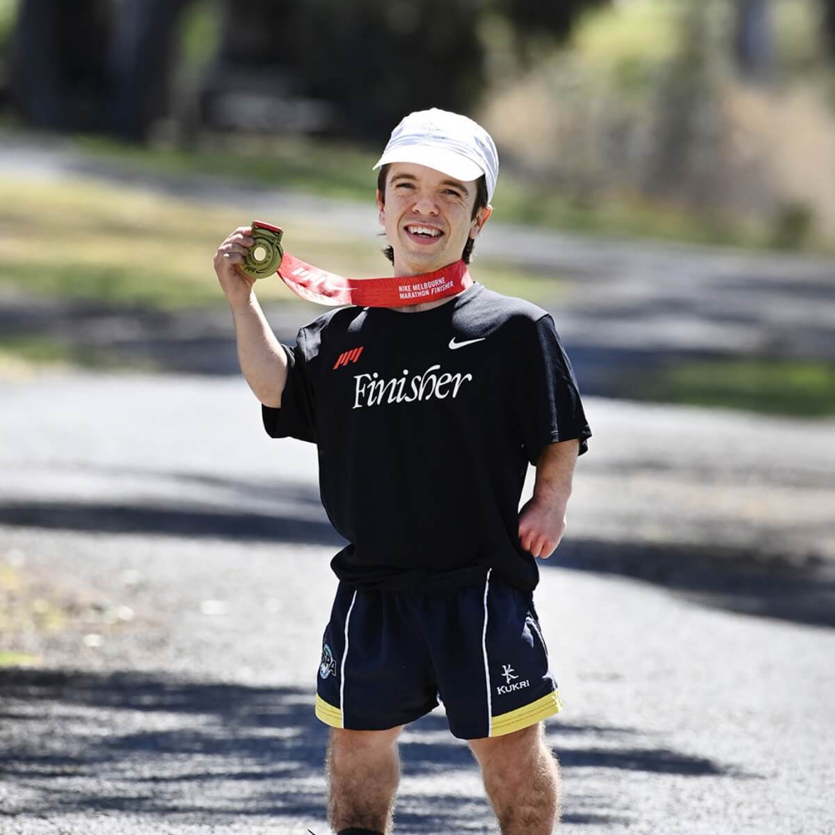 A smiling runner in a black "Finisher" shirt holds a medal, wearing a cap, standing on a path in a park.