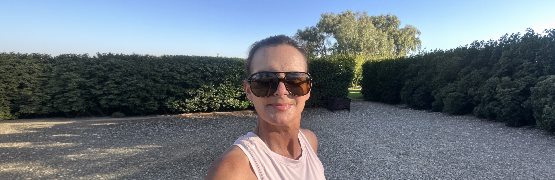 A woman in sunglasses smiles for a selfie in a gravel area surrounded by tall green hedges and trees under a clear sky.