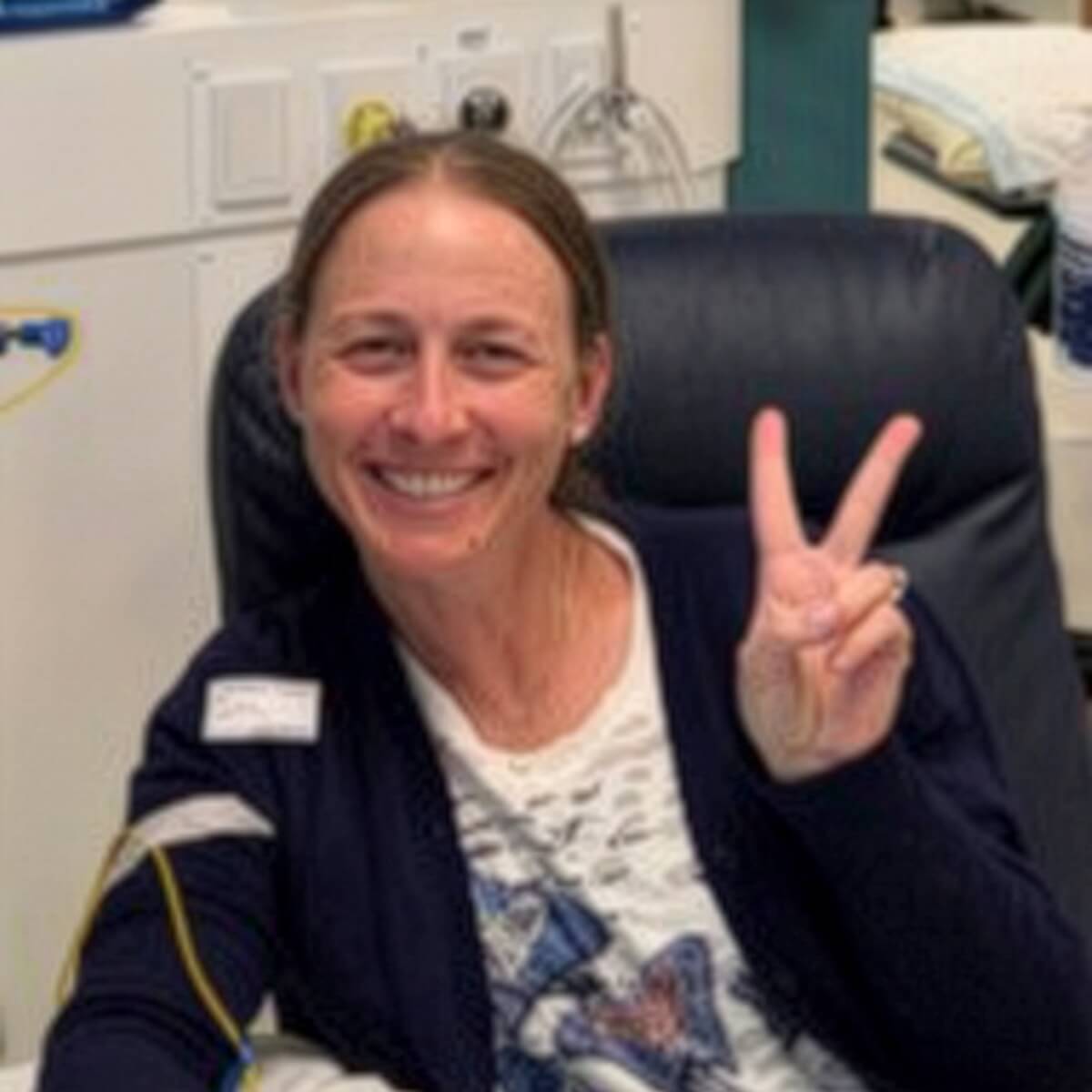 A smiling woman sitting in a chair, making a peace sign with her right hand.