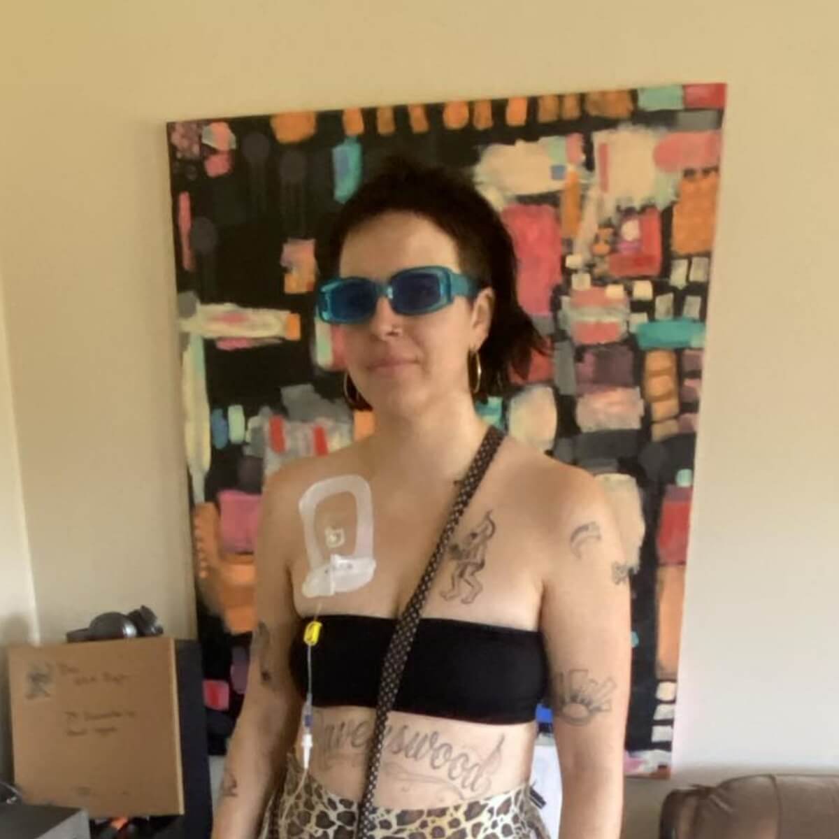 A person with tattoos, wearing a black crop top and blue sunglasses, stands in front of a colorful abstract painting.