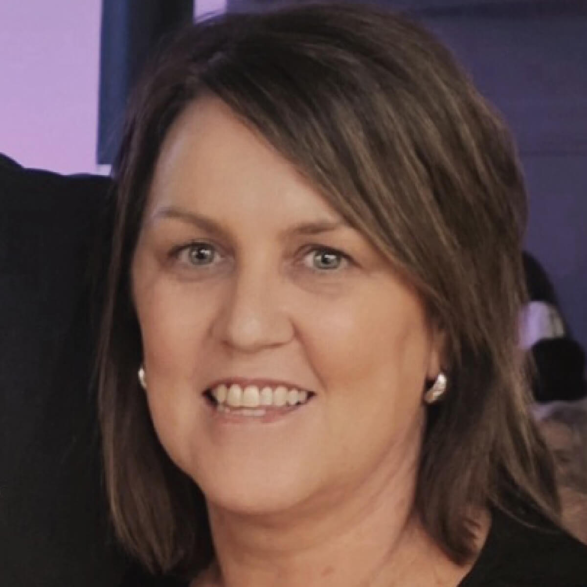 A woman with shoulder-length brown hair smiles, wearing a black top and silver earrings, against a blurred background.