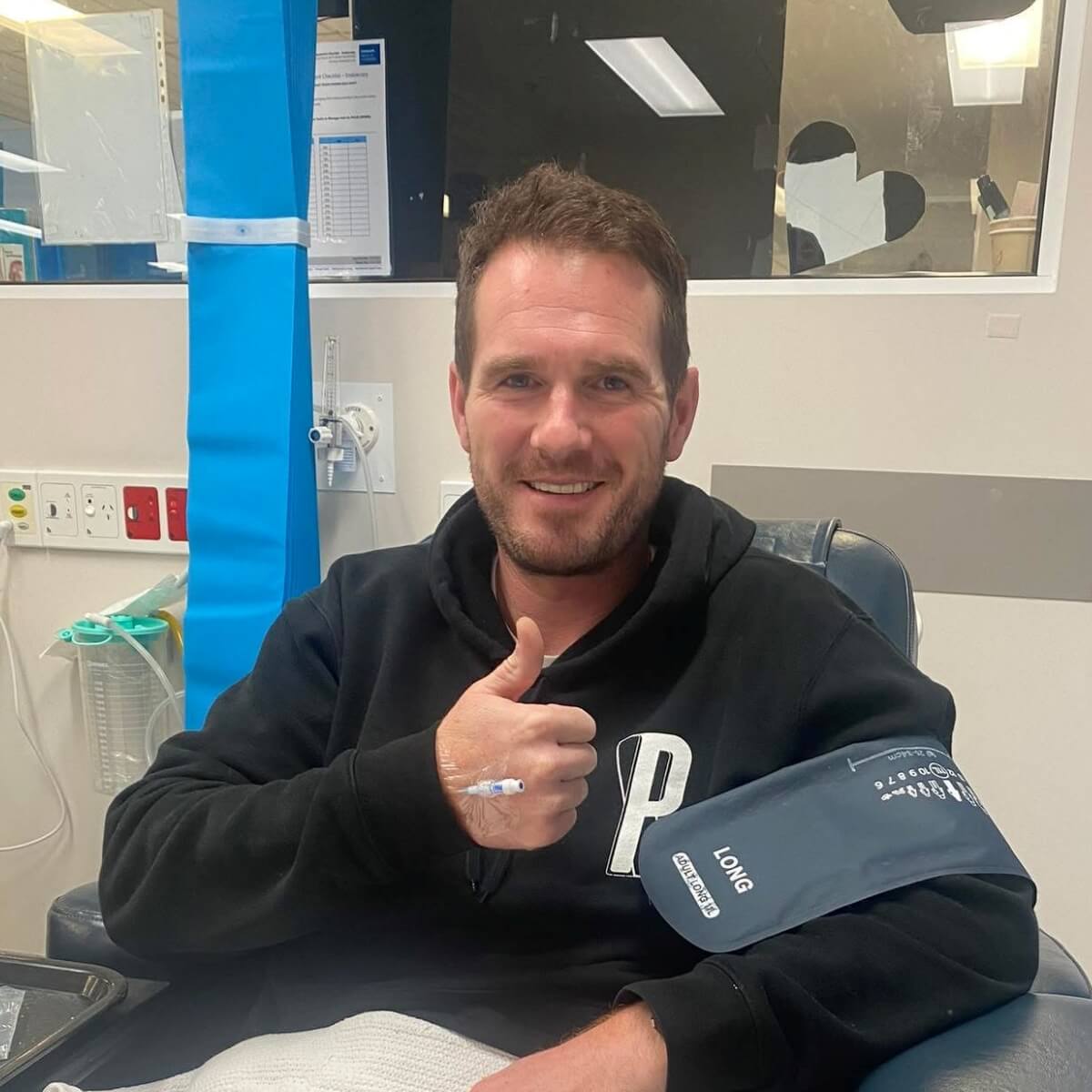 A man in a black hoodie smiles and gives a thumbs up while sitting in a medical chair with a blood pressure cuff on his arm.