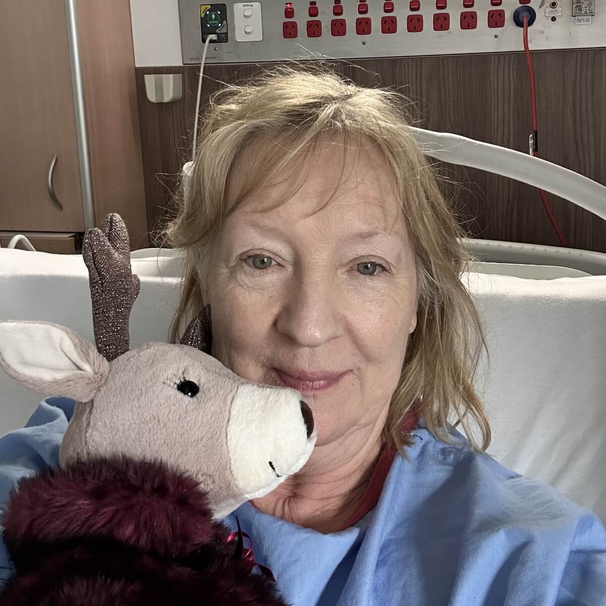 A woman in a hospital bed holds a plush toy deer, smiling at the camera.