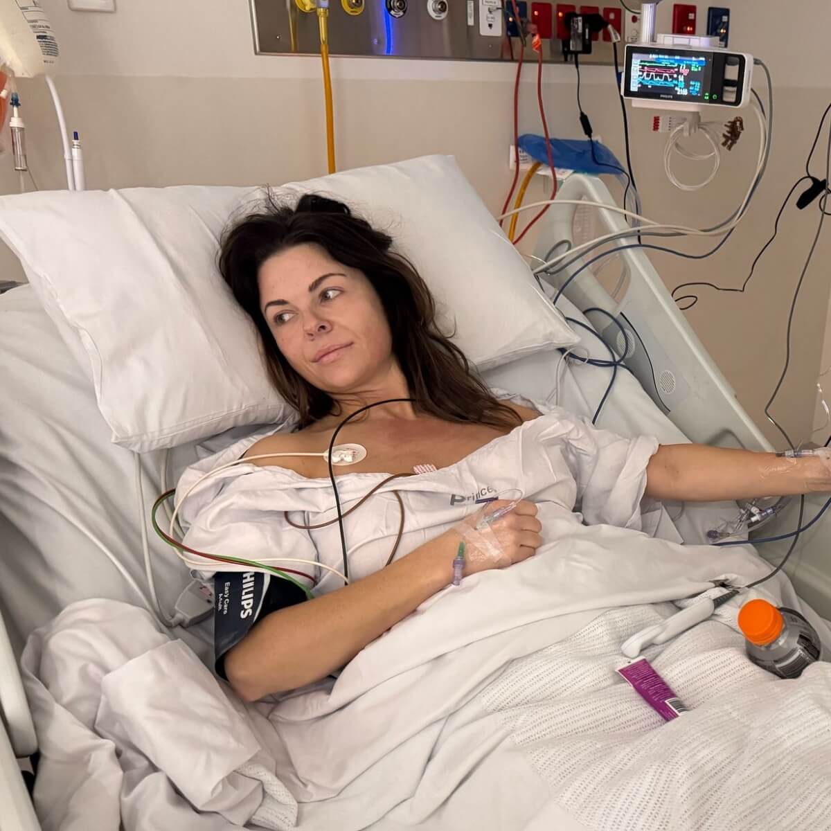 A woman in a hospital bed with medical wires and monitors, looking thoughtful while wearing a hospital gown.