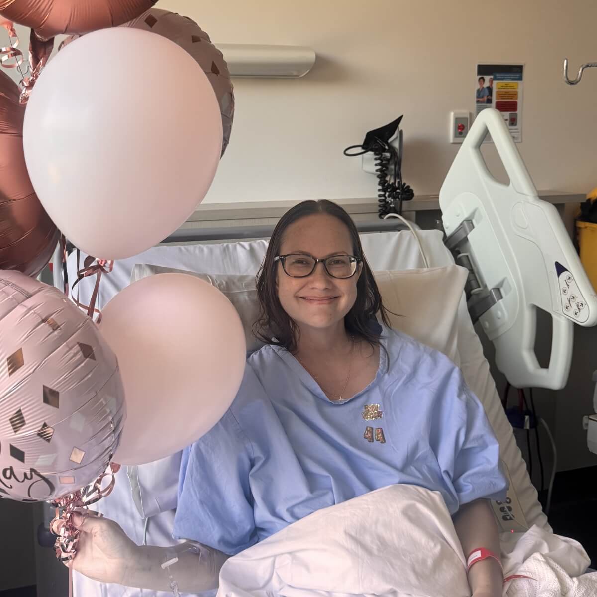 A smiling woman in a hospital bed holds balloons, wearing a blue hospital gown and glasses.