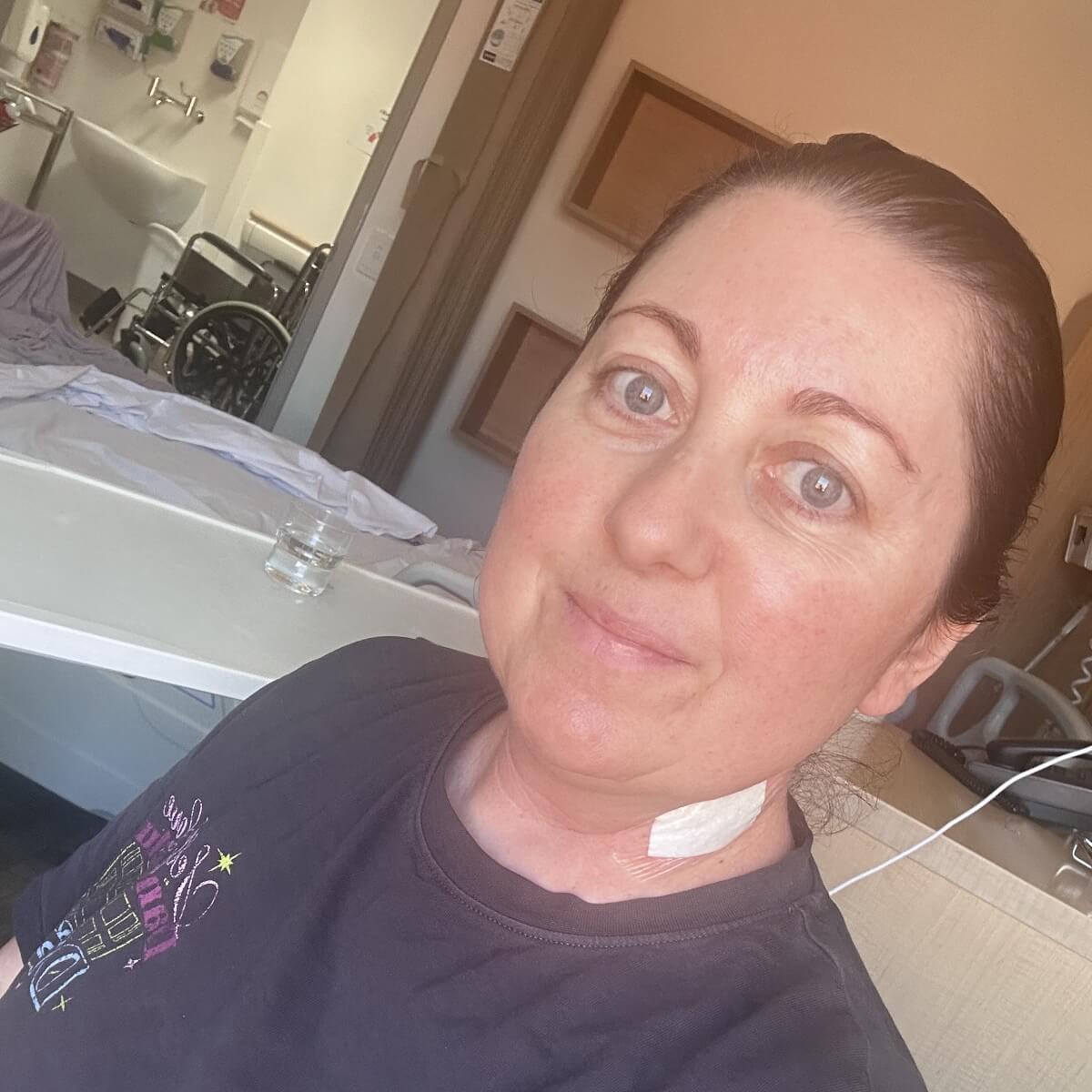 A woman with short dark hair and a bandage on her neck smiles in a hospital room with a wheelchair in the background.