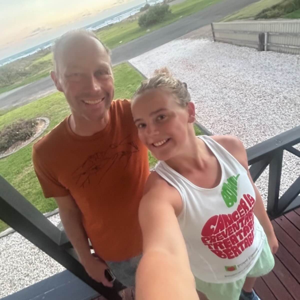 A man and a woman smile for a selfie on a porch with a grassy area and ocean in the background.