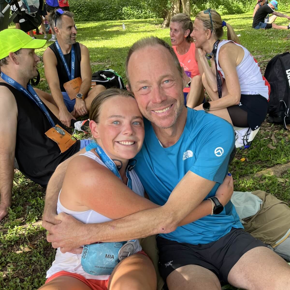 A young woman and a man sit together, smiling and embracing, with other runners resting in the background.