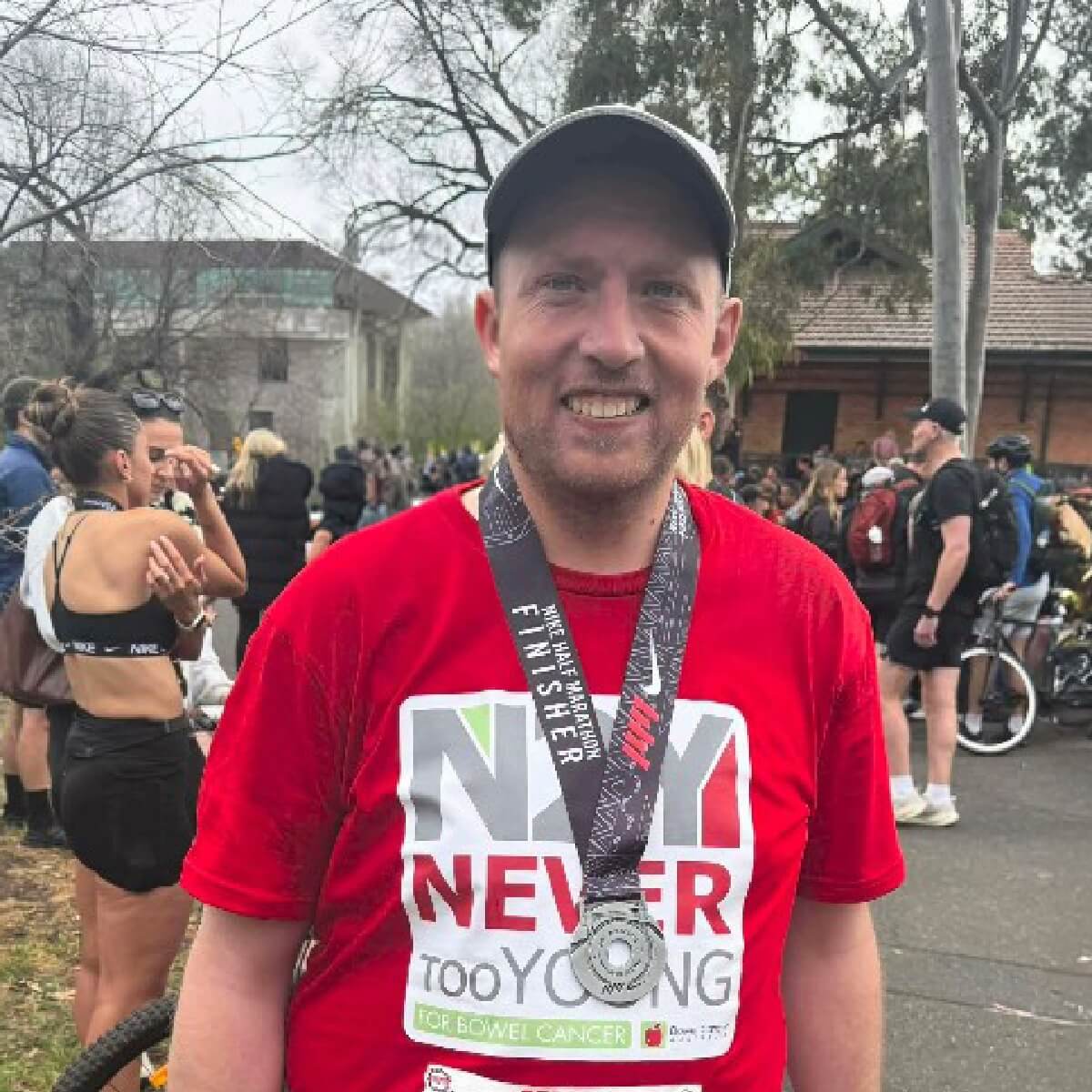 A smiling man in a red shirt with a medal stands among a crowd at an outdoor event, trees in the background.