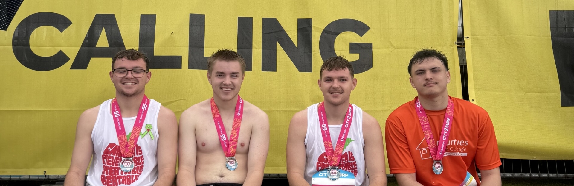 Four young men sit in front of a yellow banner that reads "CALLING," wearing medals from a race.