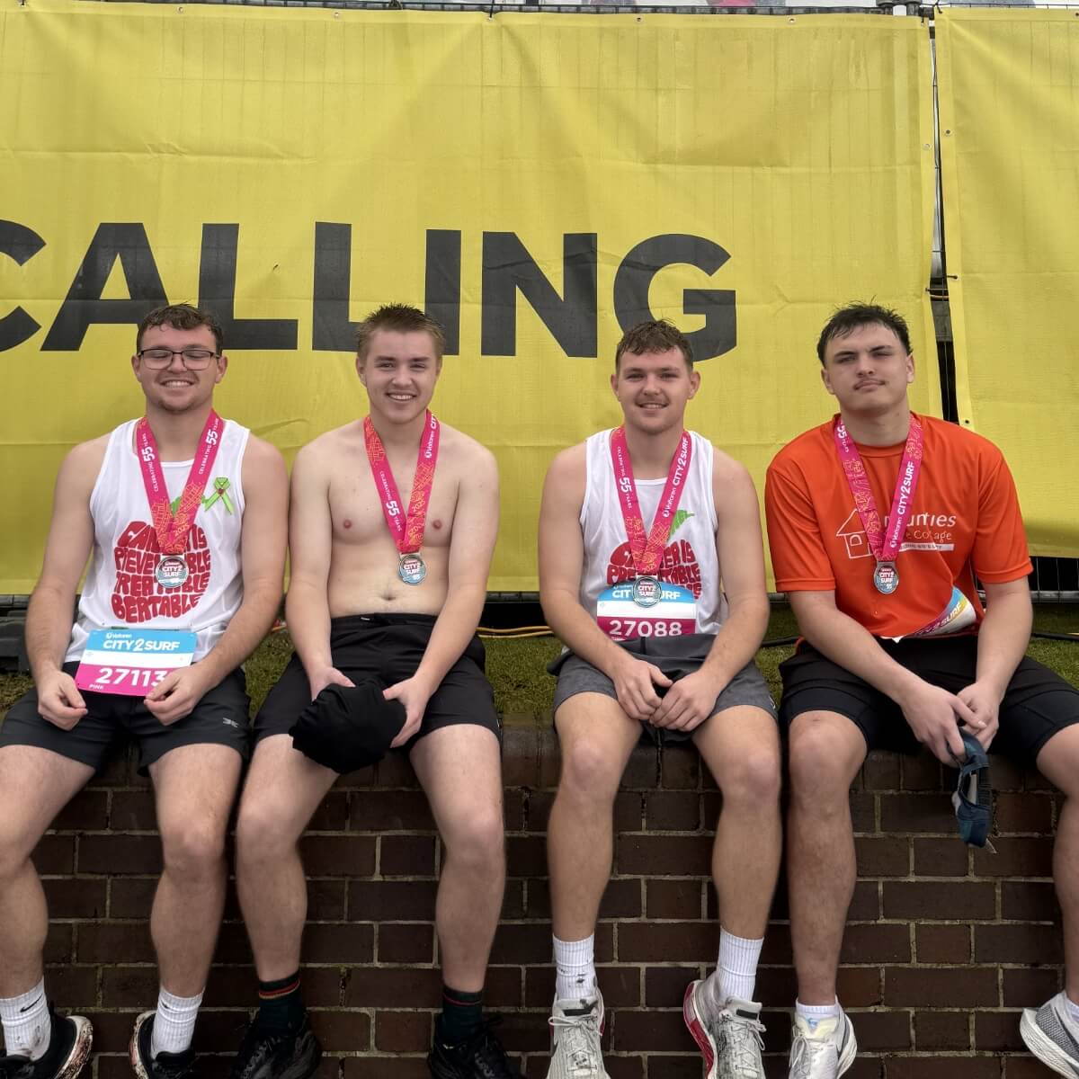Four young men sit on a brick wall, wearing medals and casual athletic clothing, in front of a yellow banner that says "CALLING."