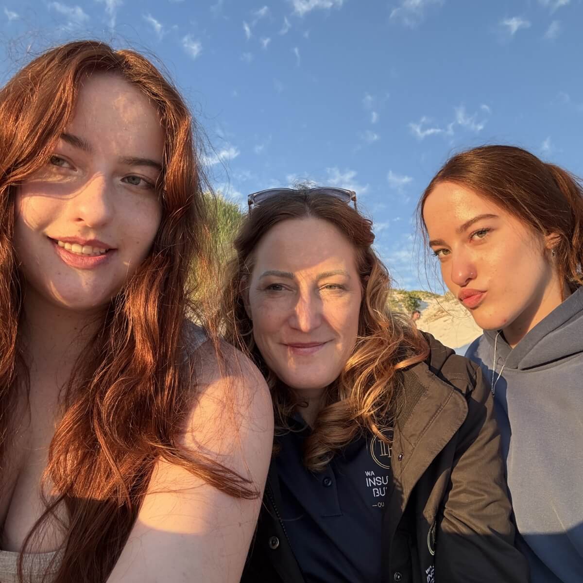 Three women with long hair smile together outdoors, with a blue sky and sandy background.