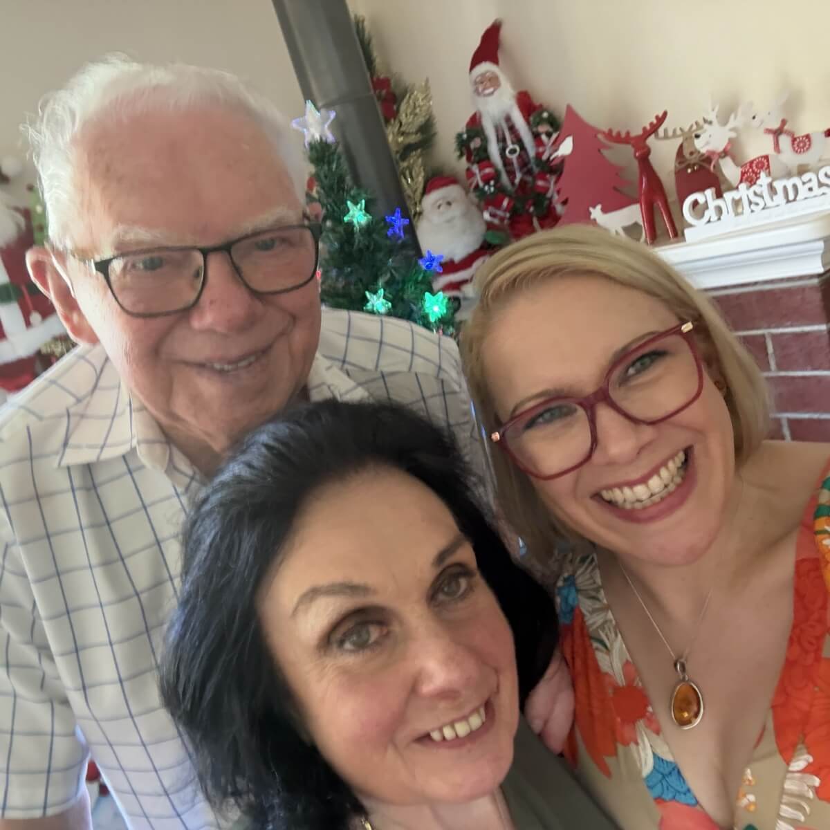 A smiling group of three people poses together in front of a festive holiday backdrop with decorations.