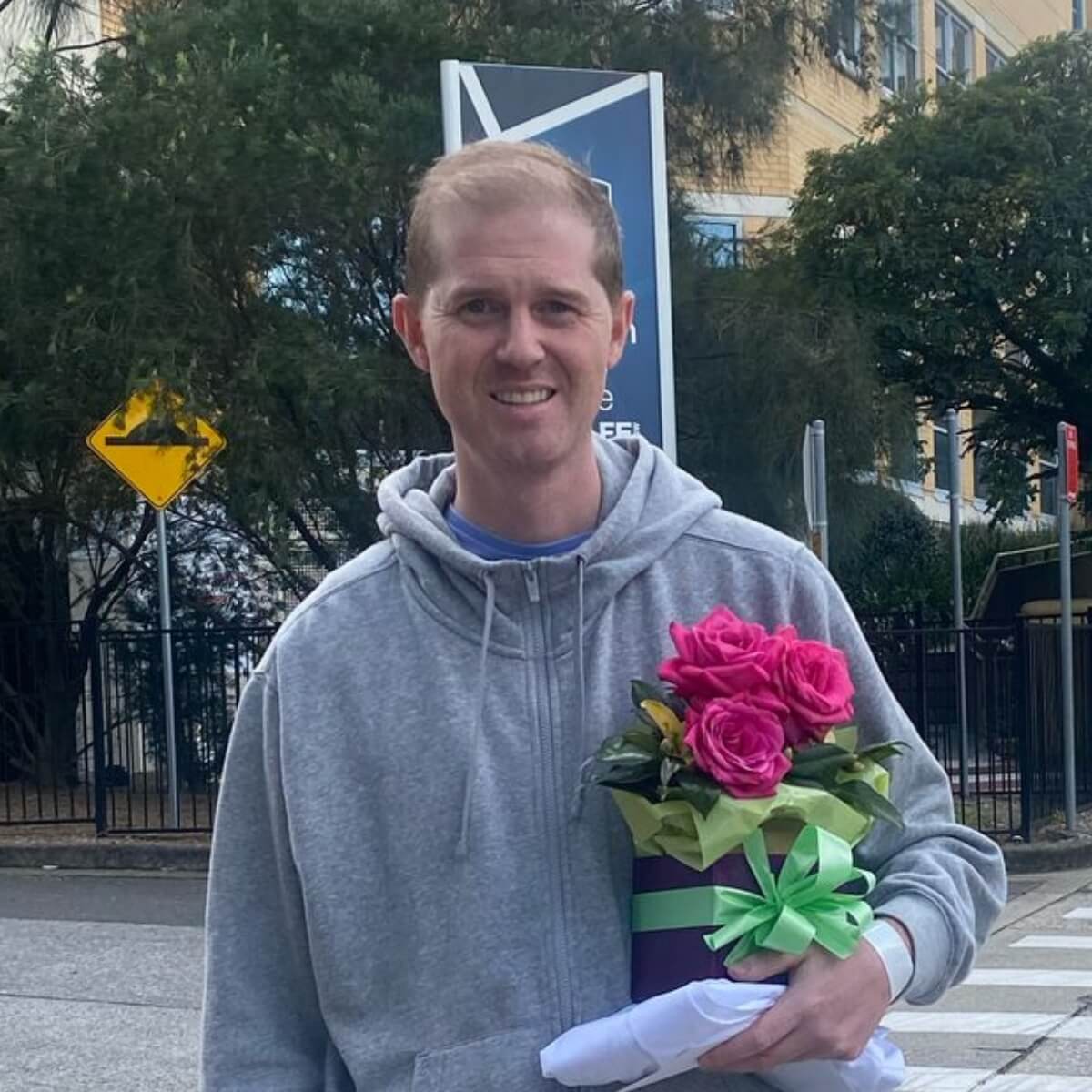 A man in a gray hoodie smiles while holding a bouquet of pink roses wrapped in green and purple.