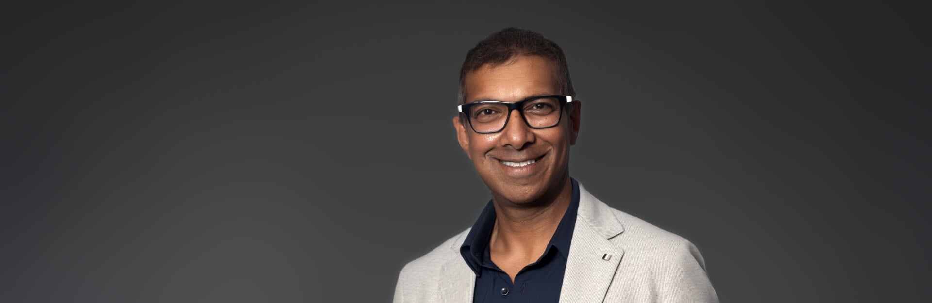 A smiling man in a light blazer and glasses against a dark gradient background.