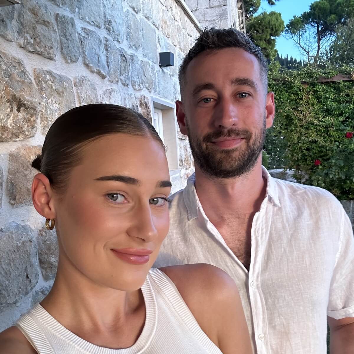 A woman with a sleek hairstyle and a man in a white shirt stand together in front of a stone wall and greenery.
