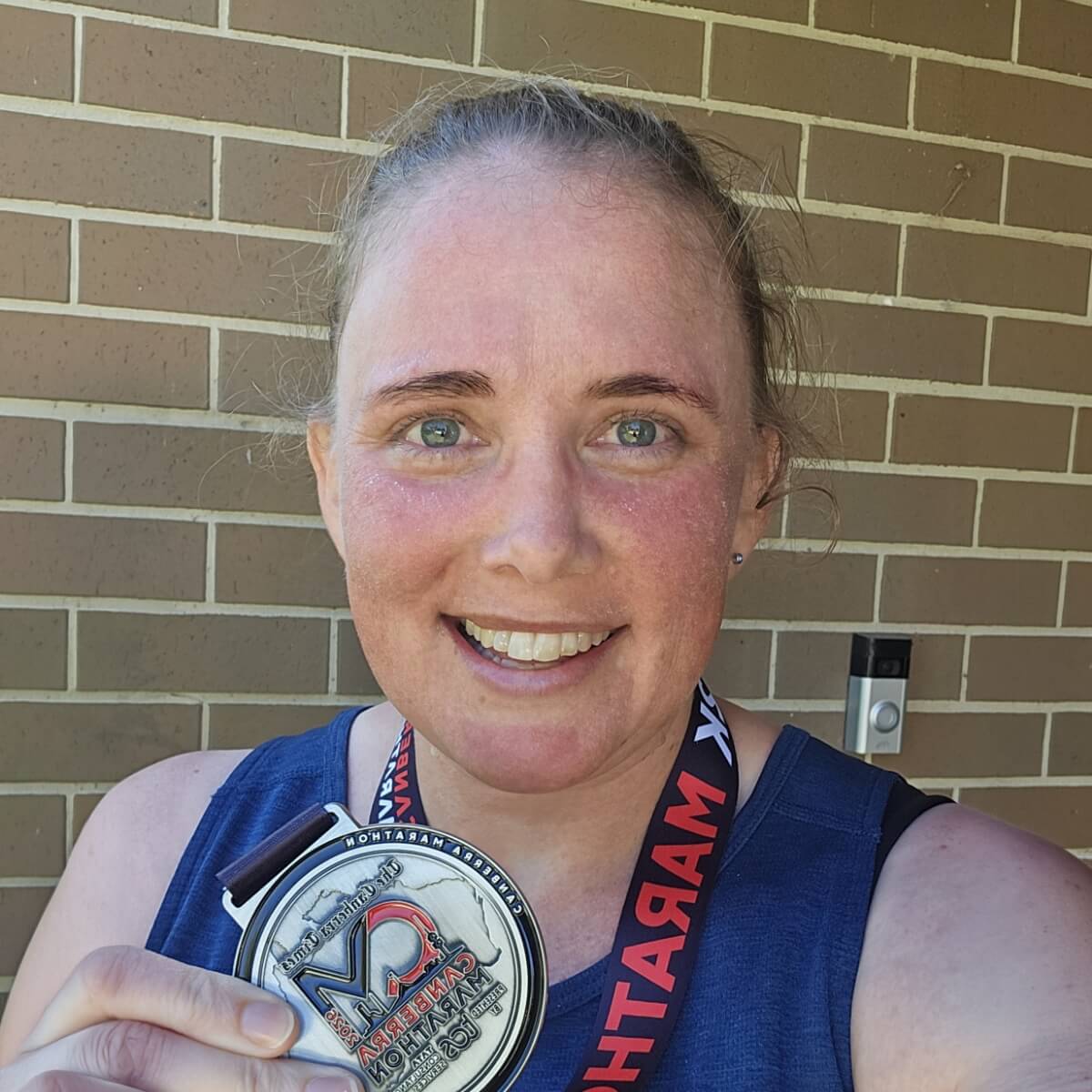 A smiling person holds a medal, standing in front of a brick wall.