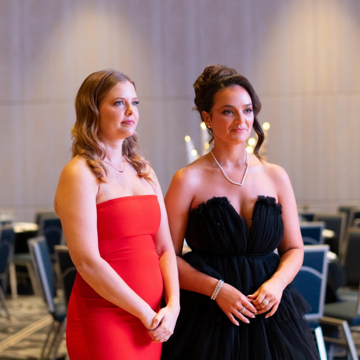 Two women stand side by side, one in a red dress and the other in a black gown, with a banquet setting in the background.