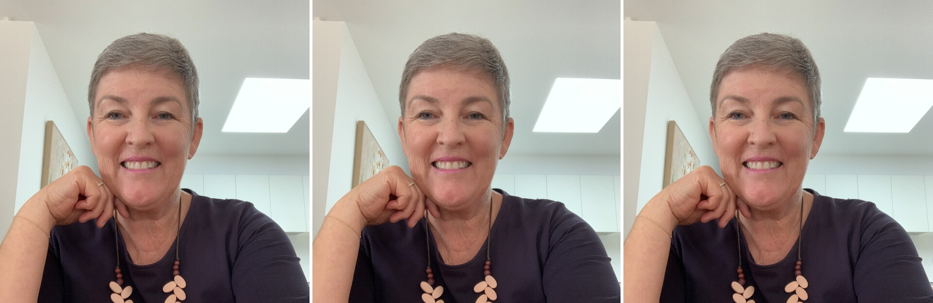 A woman with short gray hair smiles while resting her chin on her hand, wearing a dark top and a beaded necklace.