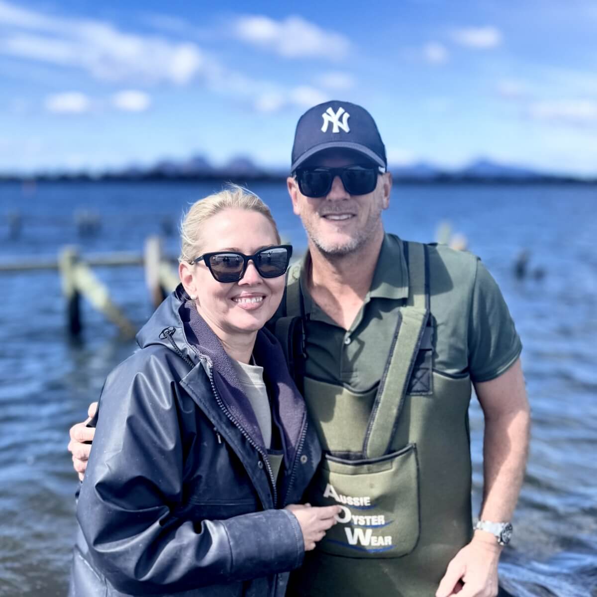 A smiling couple stands by the water, wearing sunglasses, with a scenic backdrop of blue sky and distant mountains.