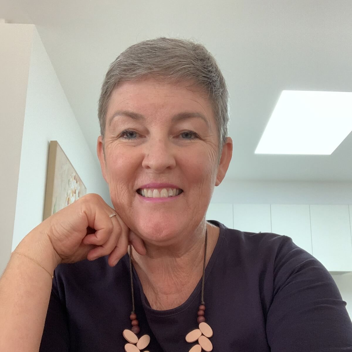 A smiling woman with short gray hair, wearing a dark shirt and a necklace, poses in a bright room.