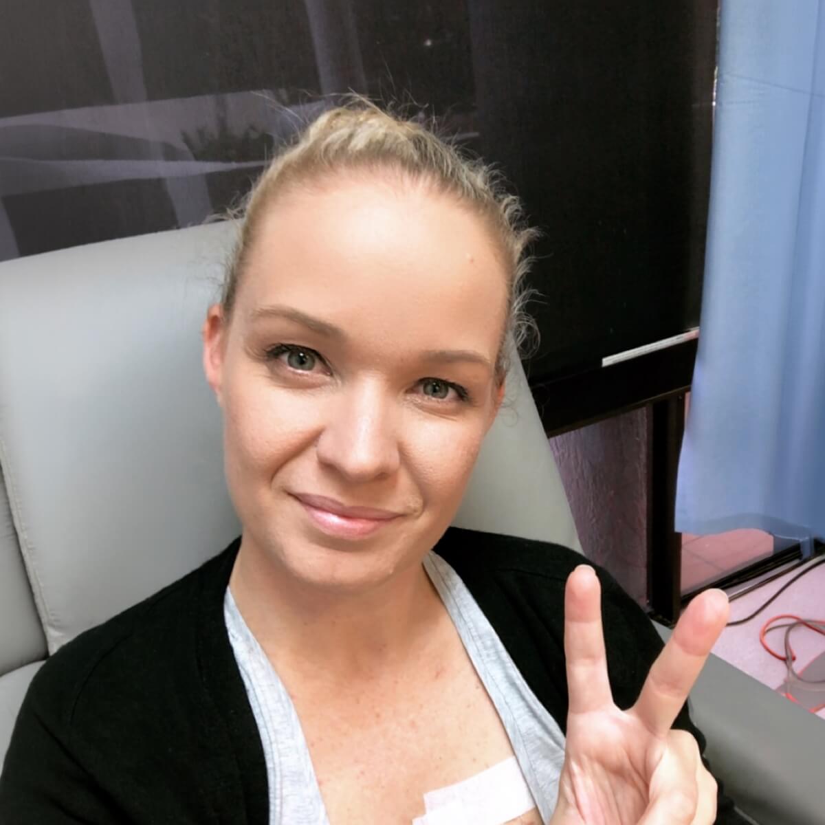 A woman with blonde hair smiles and makes a peace sign while seated in a medical setting.