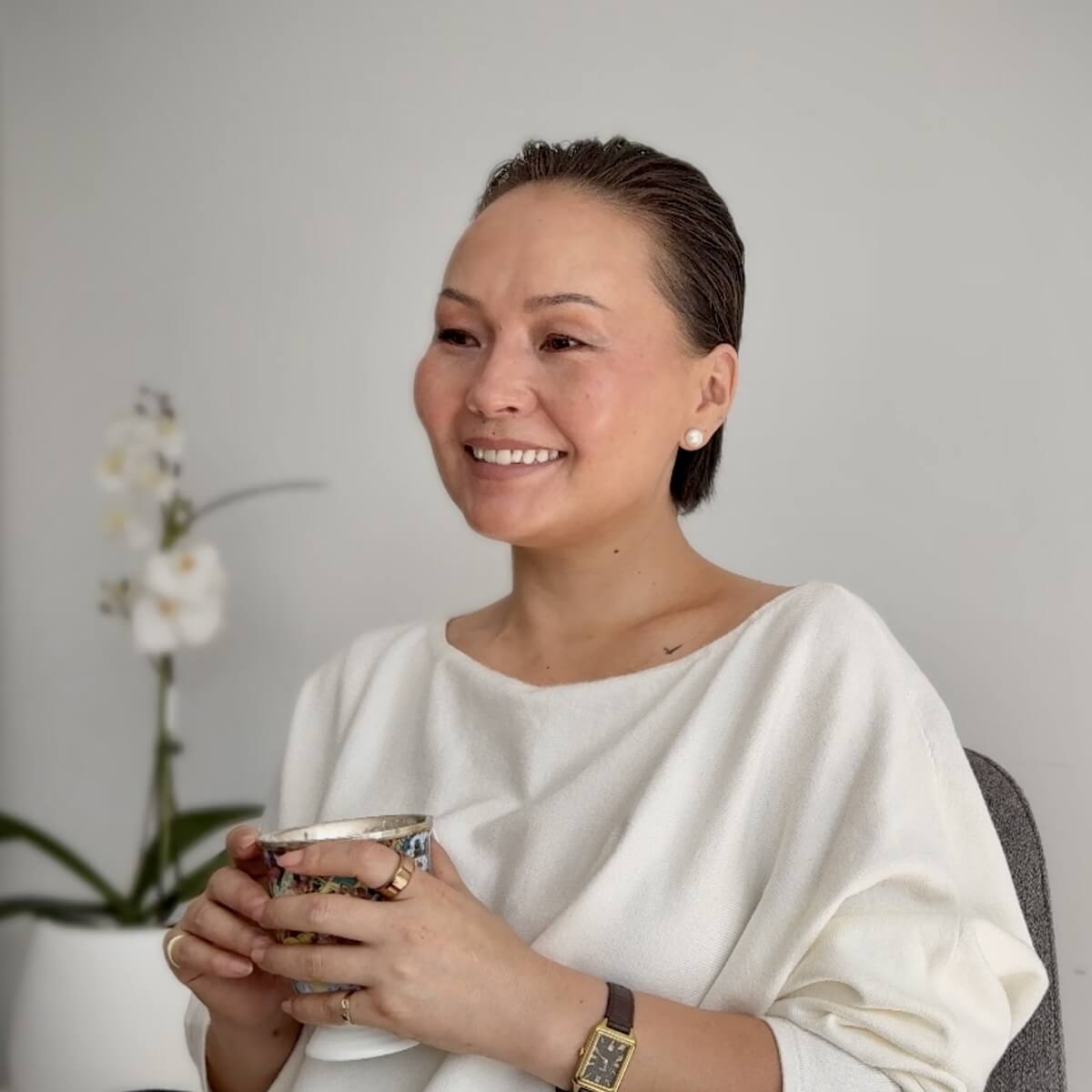 A woman with short hair smiles while holding a decorative cup, seated in a minimalist room with a plant and flower.