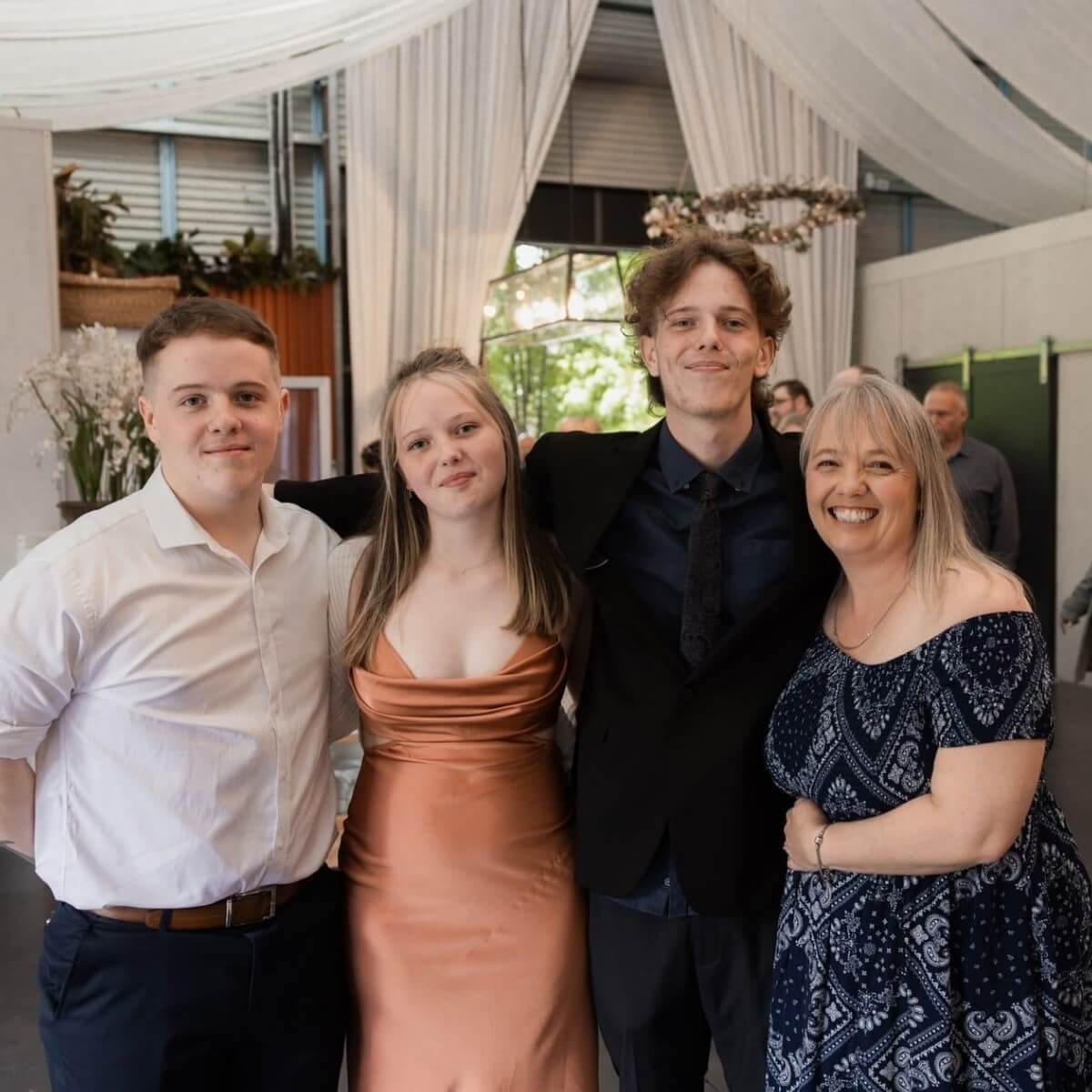 Four people pose together; two young men, a young woman in a dress, and a woman in a patterned dress, smiling.