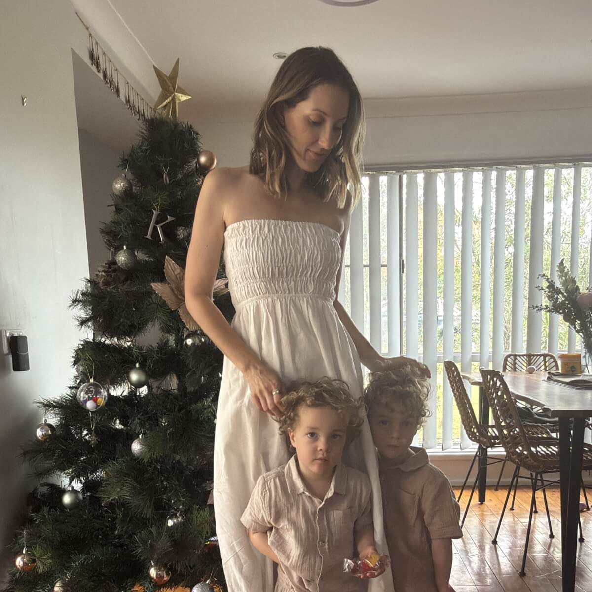A woman in a white dress stands with two young boys in front of a Christmas tree, with a dining area in the background.