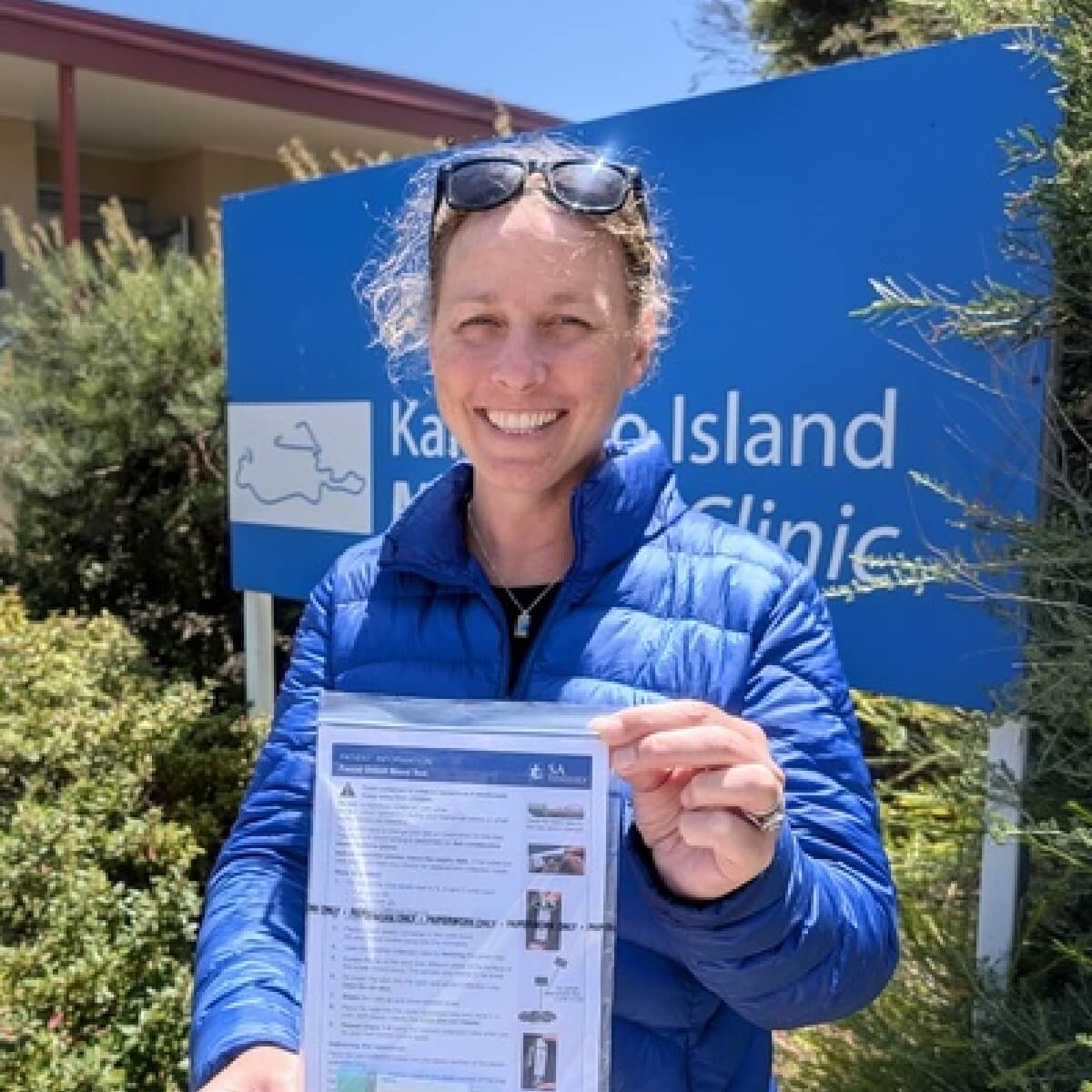 A woman in a blue jacket smiles while holding a document outside the Kangaroo Island Marine Clinic sign.
