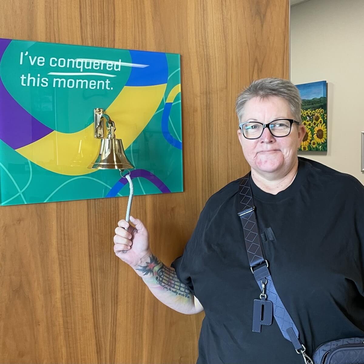 A person with short hair and glasses stands next to a bell and a sign that says, "I've conquered this moment."