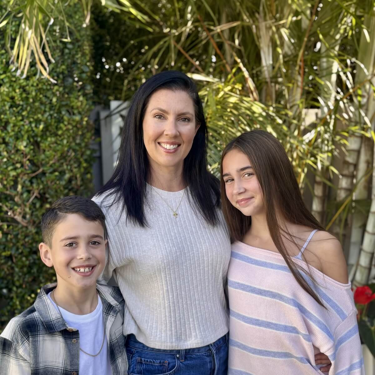 A woman stands with a boy on her left and a girl on her right, smiling, surrounded by greenery.