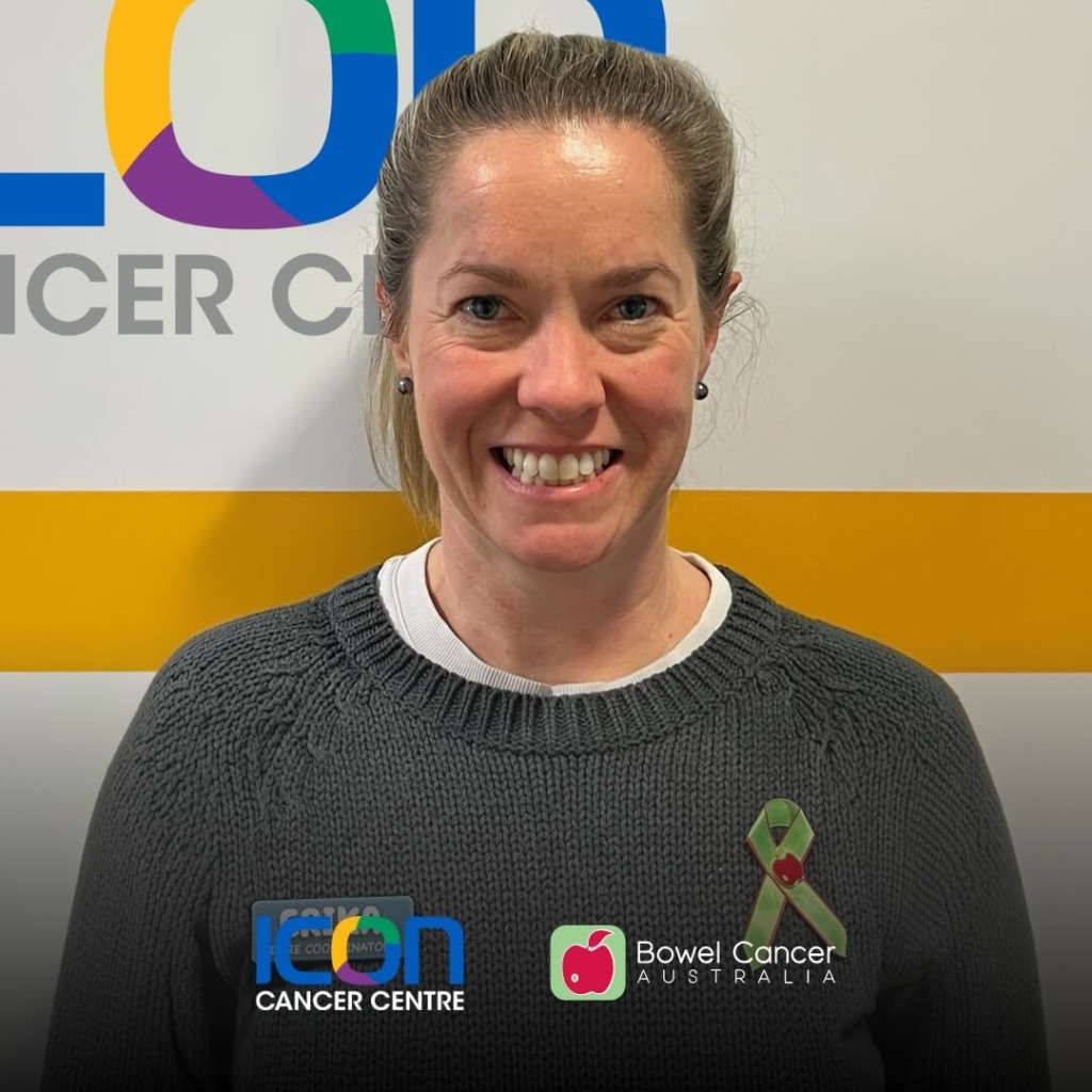 A smiling woman in a gray sweater stands in front of a colorful cancer center logo, wearing a green ribbon.
